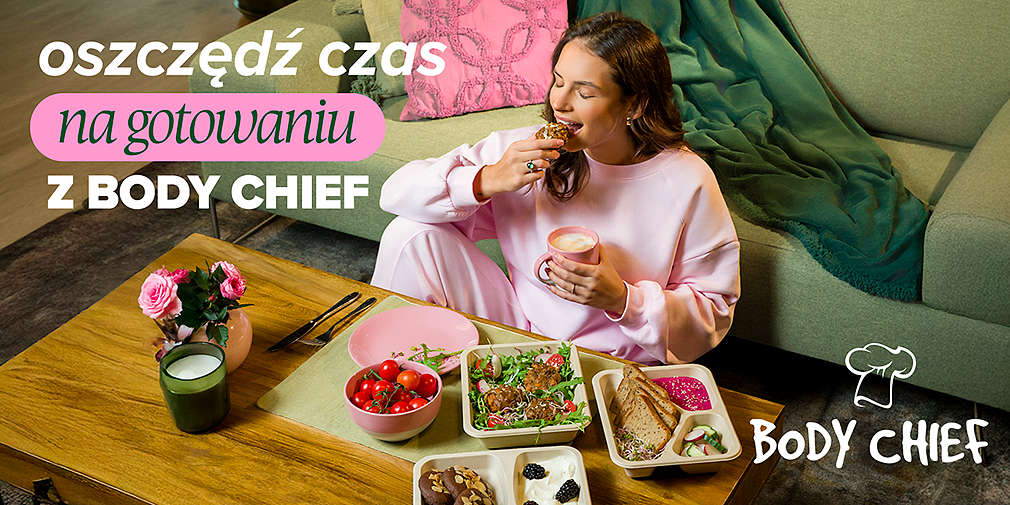 Kobieta gryzie jedzenie, siedzi na ziemi przed kanapą, przed stołem na którym jest kilka dań.