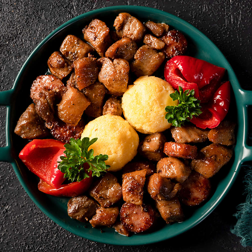 Romanian festive dish pomana porcului consisting of pieces of fried pork and sausages with pickled red peppers and mamaliga close-up in a plate on dark background. Horizontal top view from above.