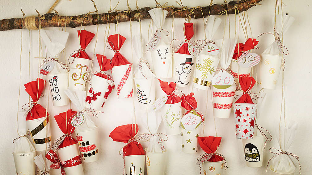 Pe o creangă groasă prinsă de un perete alb atârnă un calendar Advent pictat, din pahare de hârtie în culorile roșu și alb.
