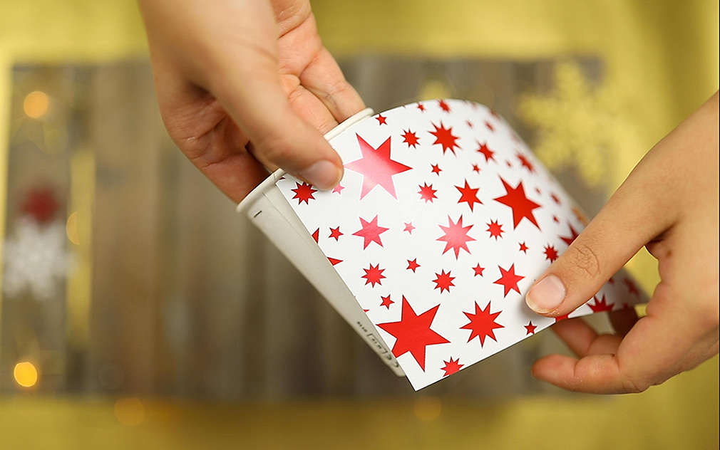 Ruce polepují bílý papírový kelímek bílým dárkovým balicím papírem s červenými hvězdami