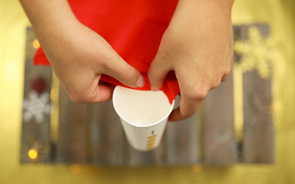 Ruce přilepují červený papír podél vnitřní strany papírového kelímku.