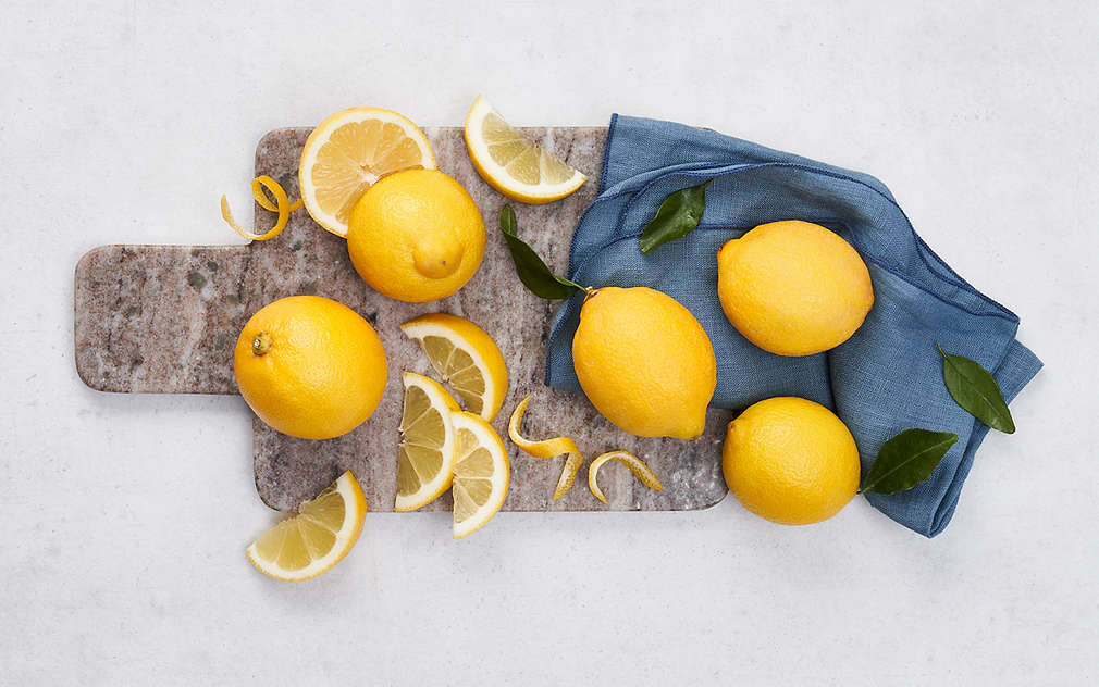 Jasne tło, kamienna deska do krojenia, częściowo zakryta niebieską ściereczką. Na desce: Cztery całe cytryny, jedna cytryna przekrojona na pół oraz plasterki i skórka cytryny.