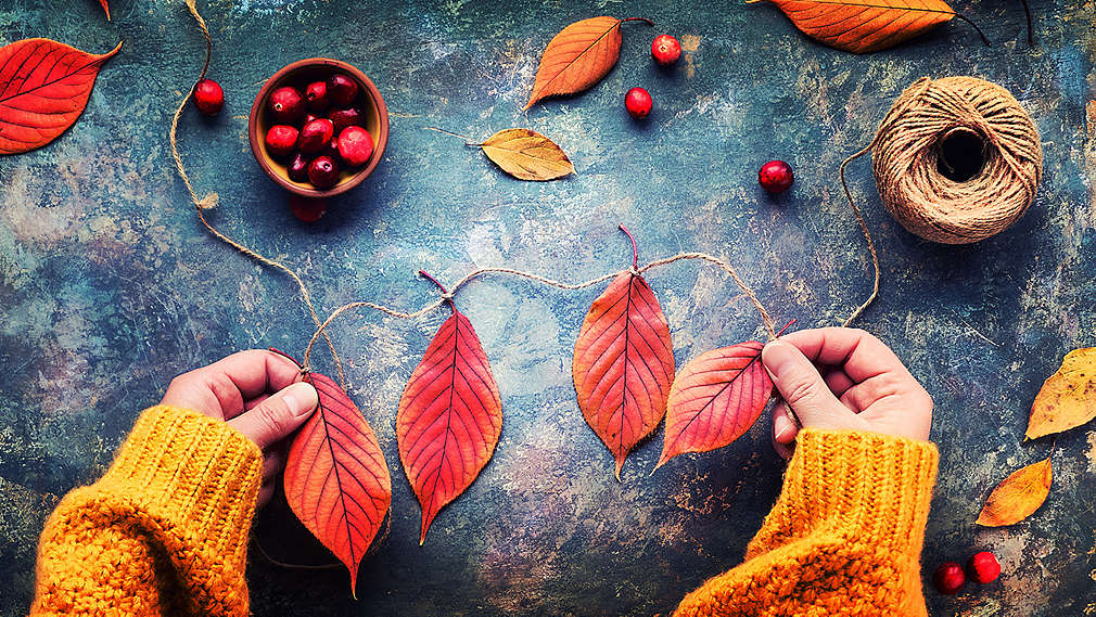 Na obrázku sú dve ruky, ktoré tvoria girlandu z červeného jesenného lístia. Podklad: modrý, v štýle batiky. Okrem toho je vidno ďalšie červené a žlté listy, červené bobule a klbko priadze.