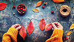 Na obrázku jsou dvě ruce, které vyrábí girlandu z červených podzimních listů. Podklad: modrý, batikovaný. Kromě toho jsou tu další červené a žluté listy, červené bobule a cívka provázku.