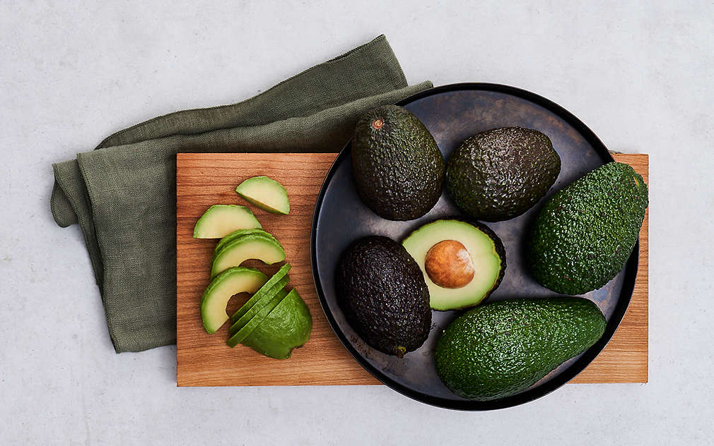 Светлосива основа, кафява кърпа, върху нея – дълга дървена дъска, върху която има фино нарязани шайби авокадо. Също така върху дървената дъска – кръгла, черна, плитка чиния, в която има пет цели авокадота и половин авокадо.