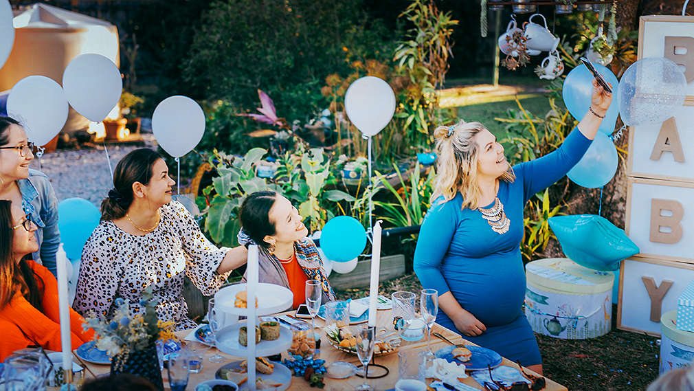 Grupa kobiet urządza piknik w ogrodzie, aby uczcić baby shower swojej ciężarnej przyjaciółki, która właśnie robi swoim gościom zdjęcie telefonem komórkowym. Kobiety siedzą wokół piknikowego koca, w tle prezenty, balony i stojak z napisem drukowanymi literami „Dziecko”.