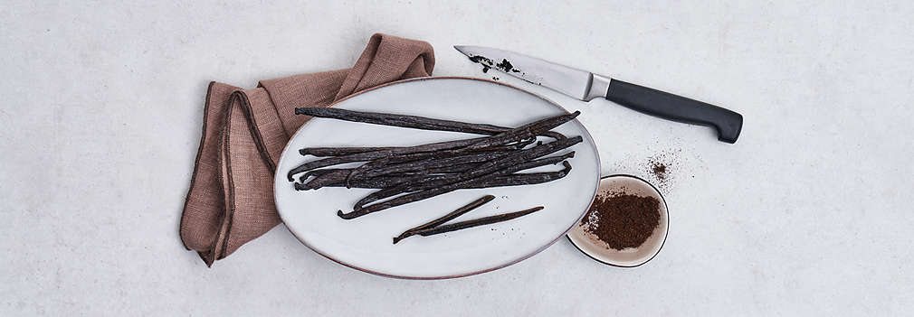 Vanilleschoten auf einem grauen Teller, darunter links ein braunes Geschirrtuch, rechts sind außerdem ein schwarzes Messer und eine kleine Schüssel mit gemahlener Vanille zu sehen