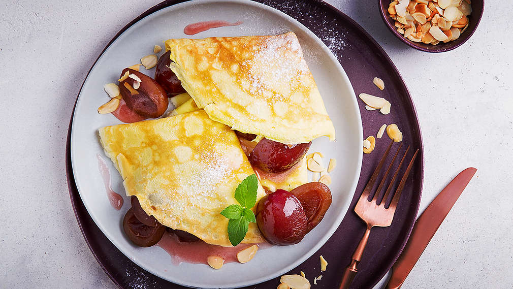 Dve na štvrtiny poskladané tenké palacinky, podávané na tanieri s omáčkou, zaváranými slivkami, medovkou a plátkami mandlí.