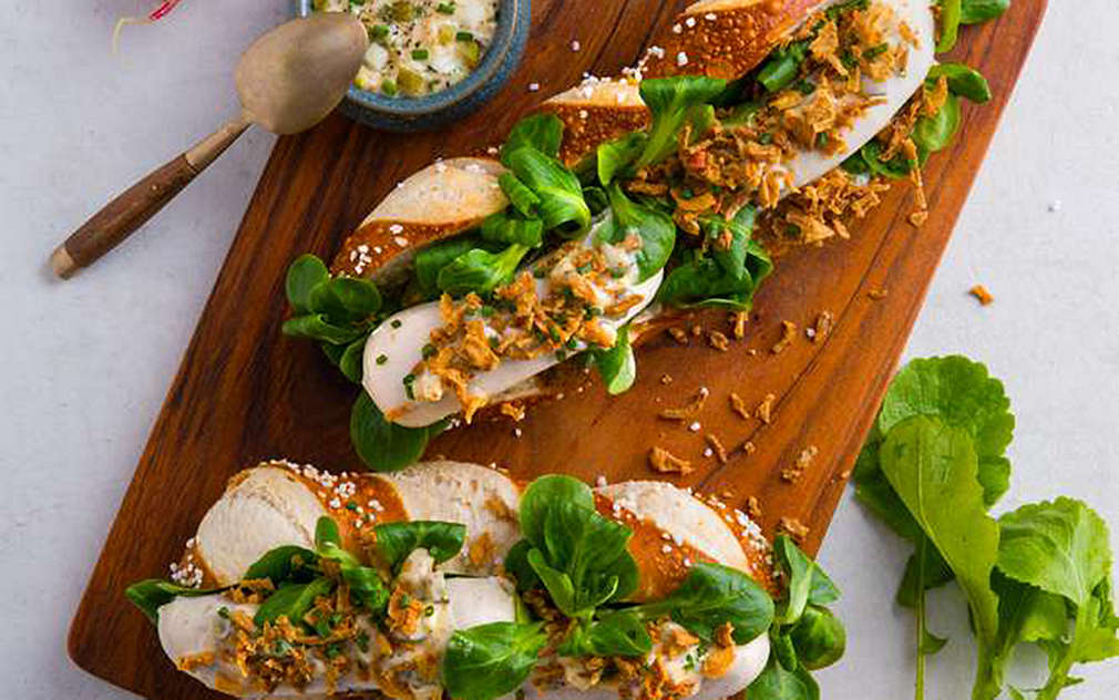 Na jasnym tle drewniana deska do krojenia z dwoma hot dogami w naciętym preclu z białą kiełbasą, roszponką, rzodkiewką. Obok stoi miseczka słodkiej musztardy.