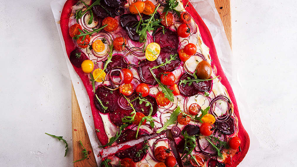 Widok z lotu ptaka na tarte flambée z czerwonego ciasta z dodatkiem buraka, pomidorków koktajlowych i krążków cebuli, na jasnej drewnianej desce.