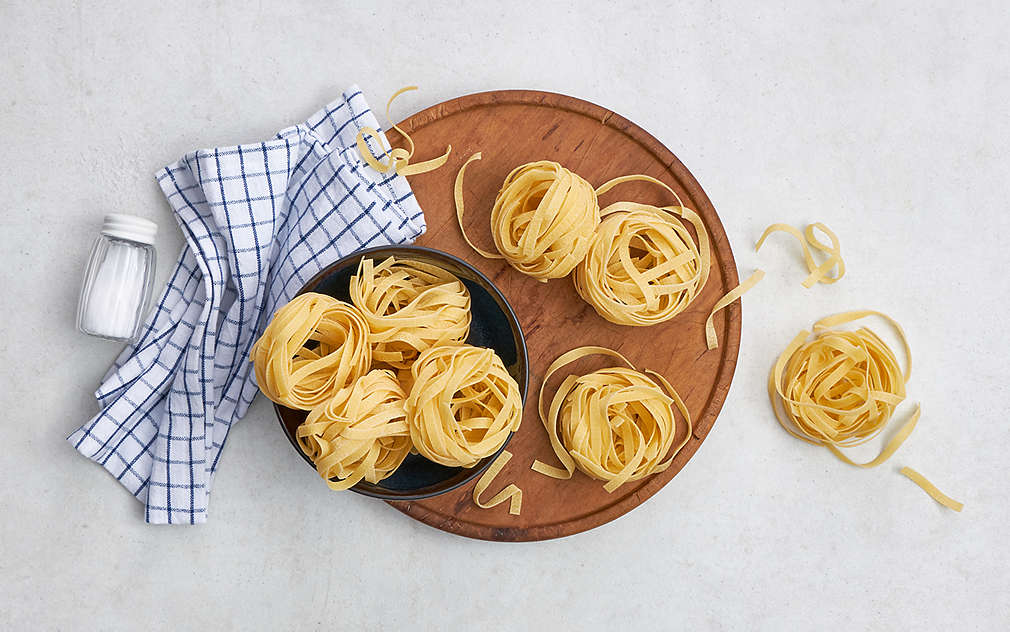 Na okrugloj drvenoj dasci nalazi se crna zdjela. I na dasci i u posudi nalaze se komadići nekuhanih tagliatella. Pokraj njih nalazi se plava karirana kuhinjska krpa i soljenka.