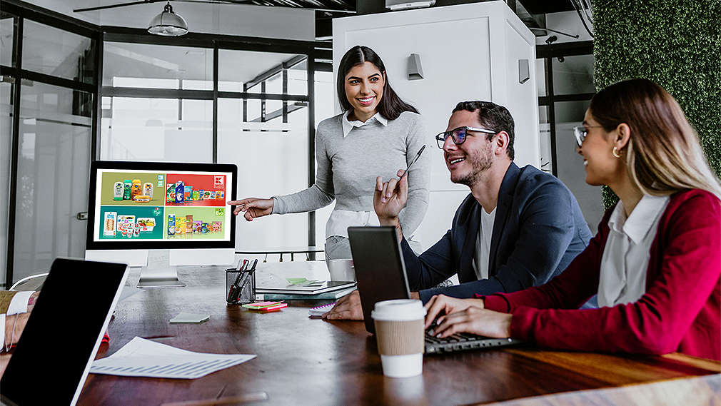 four people sit at a table and discuss something together. A woman stands in front of a screen and points to various private label collages