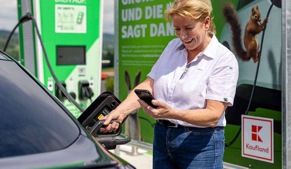 Abbildung: Eine Frau steht mit ihrem Smartphone an einer E-Ladesäule von Kaufland