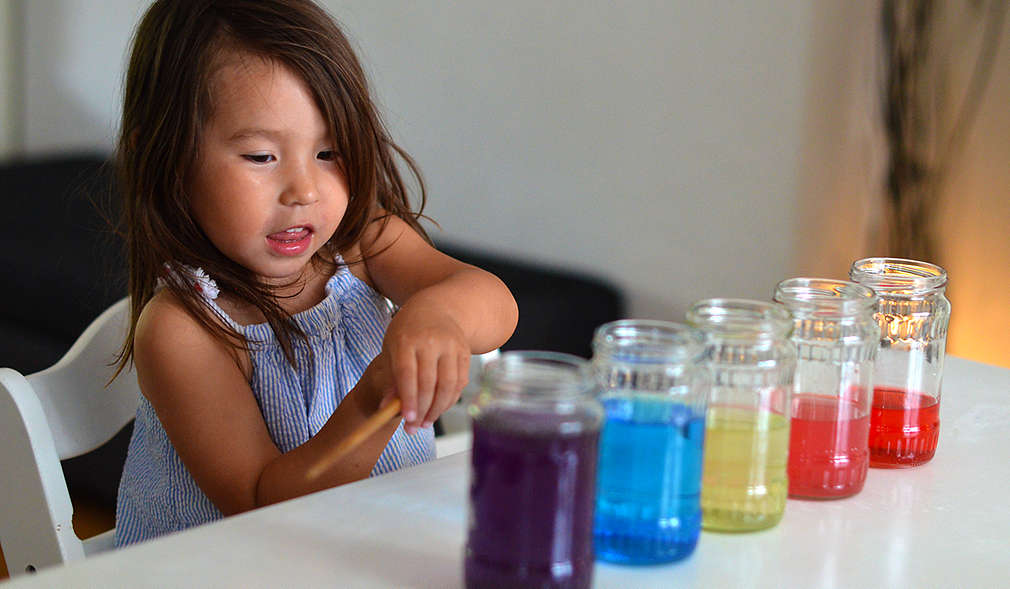 Kleines Mädchen sitzt am Tisch, darauf stehen Gläser mit bunten Flüssigkeiten befüllt
