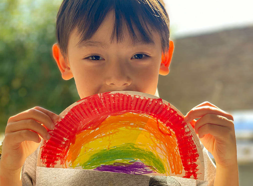 Junge hält einen aus Pappe, Schnipseln und Buntstiften gebastelten Regenbogen