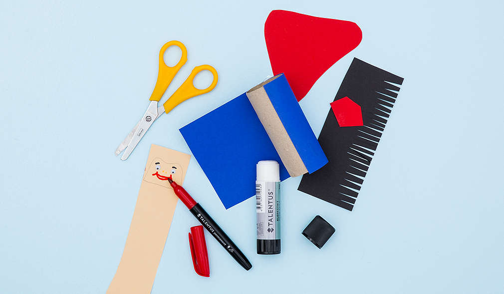 Schwarze, eingeschnittene Pappe; gelbe Schere; gemaltes Gesicht auf beigem Papier; offener Klebestift; in blaue Pappe eingewickelte Klopapierrolle, Umhang aus roter Pappe