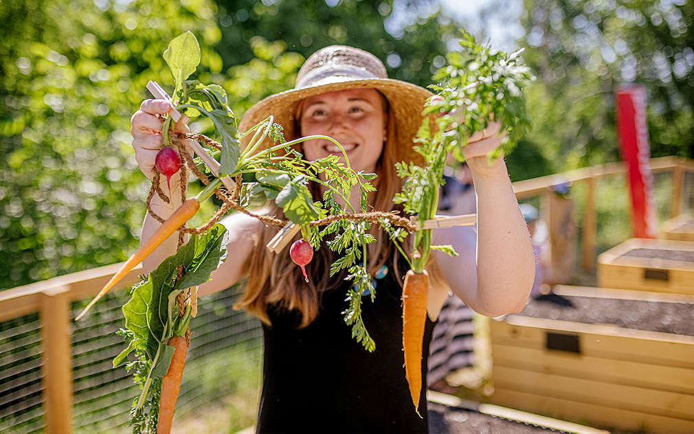 Mladá žena v klobouku drží ředkvičky a mrkev.