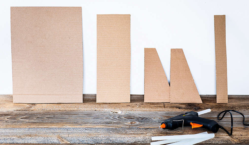 Auf einer rustikalen Unterfläche aus Holz sind Pappteile in unterschiedlichen Formaten an eine weiße Wand gelehnt. Auf der Unterfläche liegt eine Klebepistole und ein Zollstock.