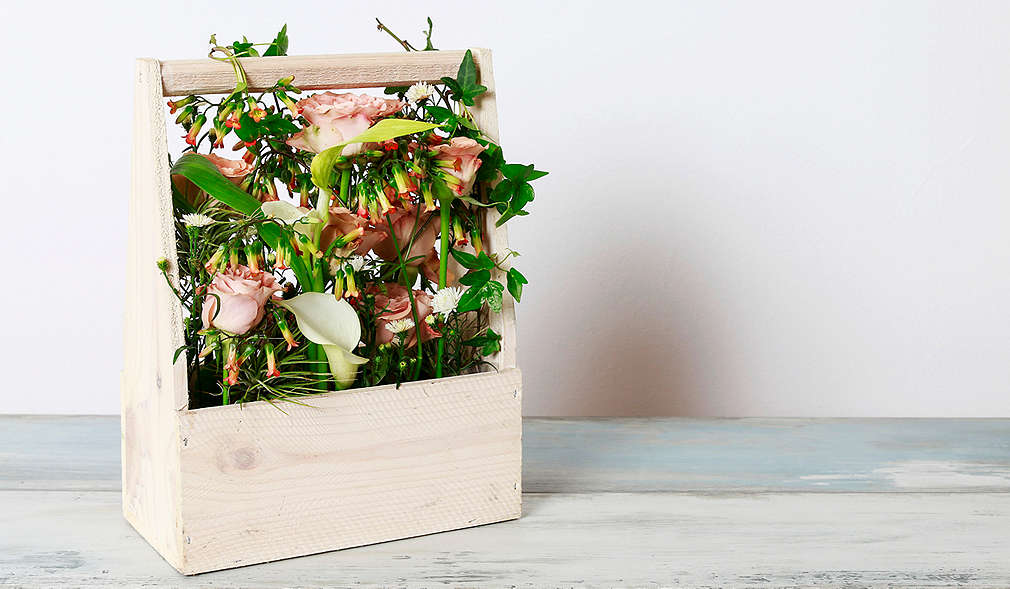 Weiß lackierte Holzfläche, darauf ein Flaschenträger, der mit frühlingshaften Blumen gefüllt ist.