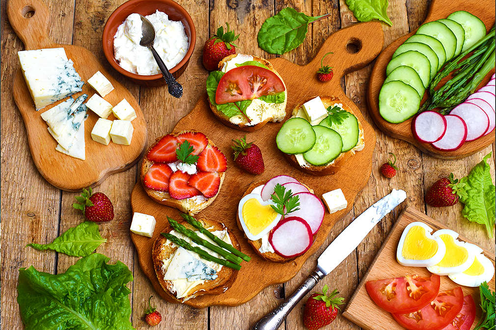 Mehrere Holzbretter liegen auf einem Holztisch. Auf den Brettern liegt aufgeschnittenes Gemüse sowie belegte Brote