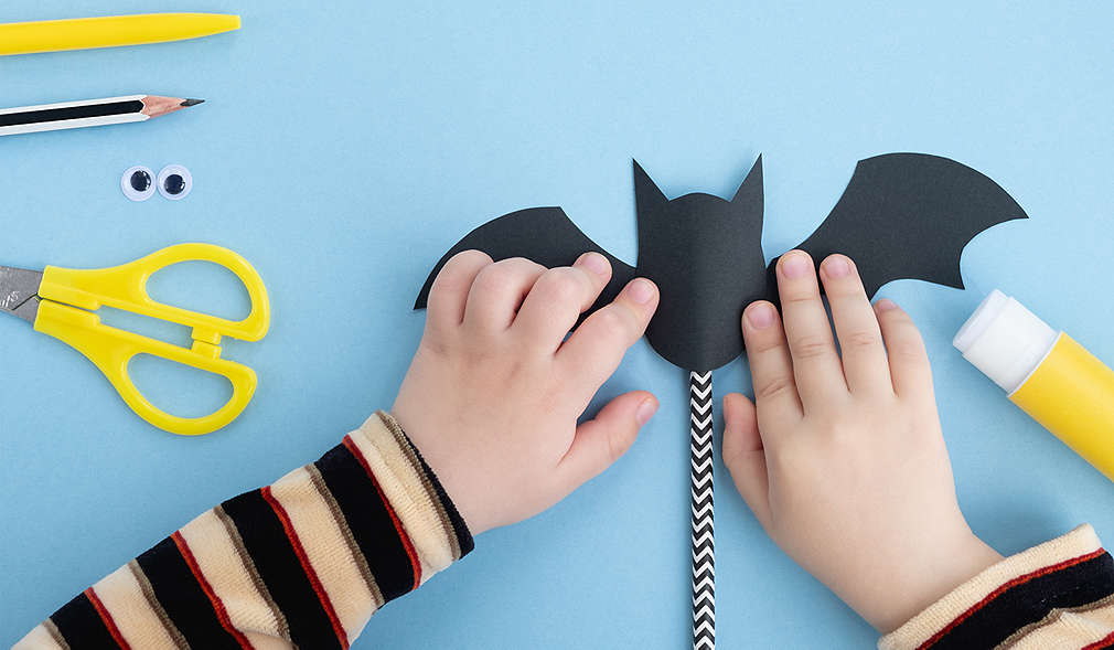 Blauer Untergrund, darauf gelbe Bastelutensilien (Stift, Schere, Kleber) und ein schwarz-weißer Bleistift, in der Mitte des Bildes basteln zwei Kinderhände eine schwarze Fledermaus aus Papier, die an einem Stab befestigt ist.