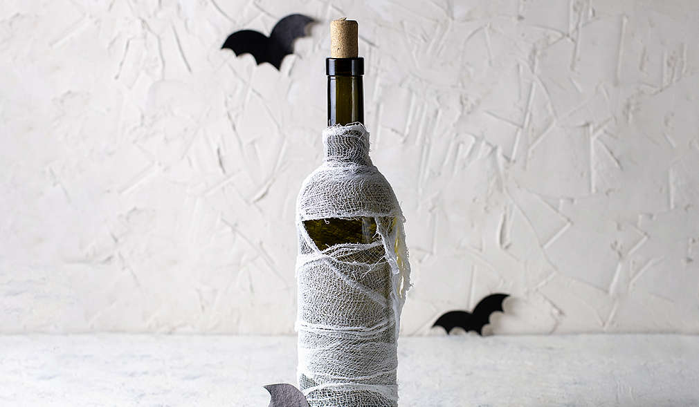 Weiße Wand mit Struktur im Hintergrund, hellgrauer Untergrund, darauf eine Weinflasche mit Korken, die mit Mullbinde umwickelt ist. Als Dekoelemente sind außerdem drei kleine Feldermäuse aus Papier im Bild.