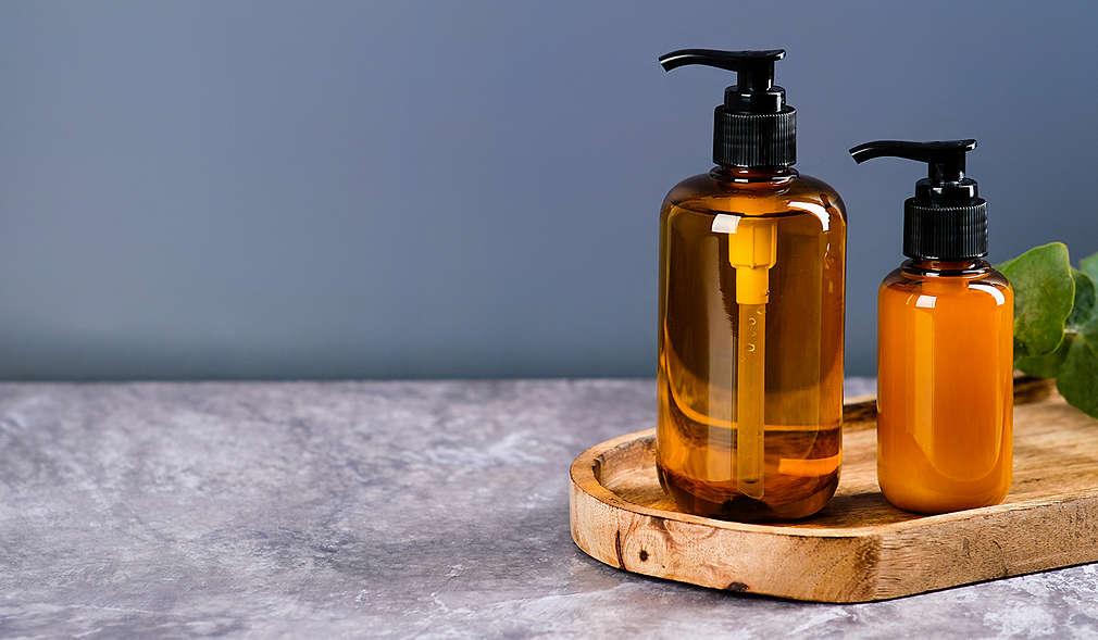 Untergrund aus grauem Marmor, darauf ein ovales Holzbrett, auf dem zwei braune Seifenspender stehen, die mit durchsichtiger und milchiger Seife gefüllt sind, graublauer Hintergrund.