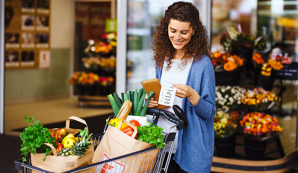 Zu sehen ist eine Frau mit vollem Einkaufswagen, die gerade aus einer Kaufland-Filiale kommt und nochmal ihre Einkaufsliste checkt.