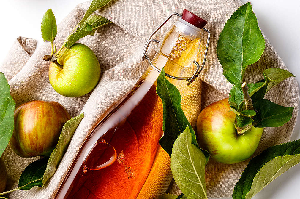 Auf einem beigen Leinentuch liegen eine Flasche Apfelessig und Äpfel mit Blättern