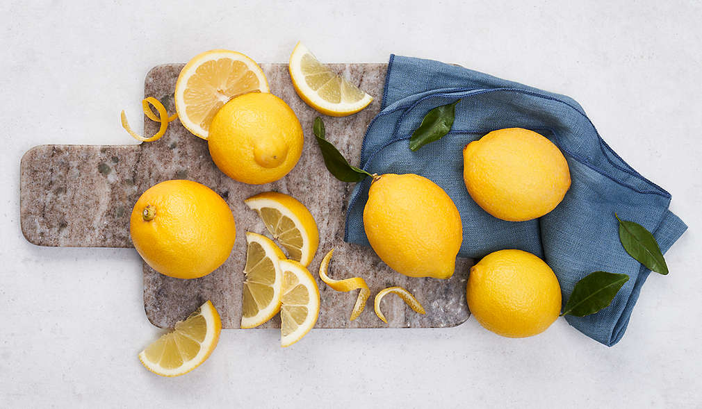 Heller Untergrund, Steinbrettchen, teilweise bedeckt mit blauem Geschirrtuch. Auf dem Brett: Vier ganze Zitronen, eine halbierte Zitrone, außerdem Zitronenscheiben und -zesten.