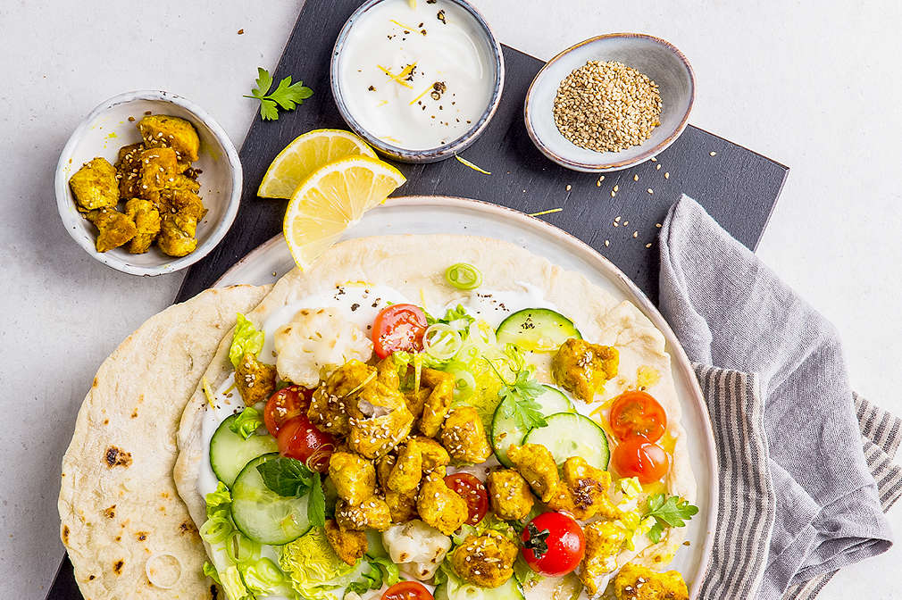 Zu sehen ist ein Teller auf einem schwarzen Brett, auf dem Teller sind Fladen, belegt mit Dip, Salatgurkenscheiben, Cherrytomaten, Salat und mariniertem und gegrilltem Tofu; seitlich des Tellers ist ein blau-weiß-gestreiftes Küchentuch zu sehen, über dem Teller sind drei Schälchen mit mariniertem Tofu, Dip und Sesamkörnern sowie zwei Zitronenscheiben zu sehen