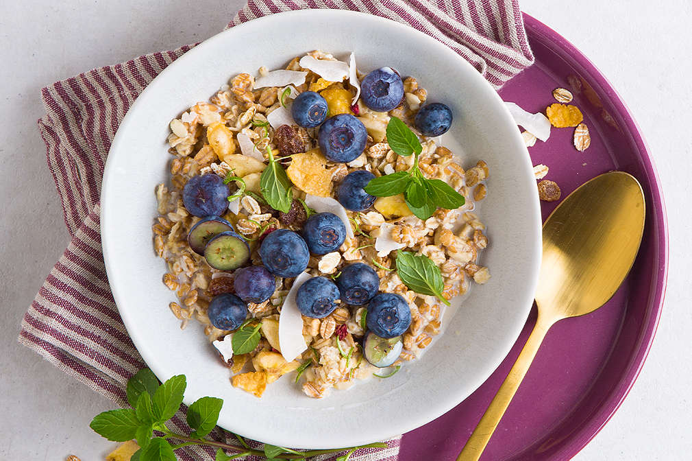 Eine Schale mit Müsli, getoppt mit Blaubeeren und Minzeblättern. Daneben ein goldener Löffel. Darunter ein brombeerfarbenes Tablett und ein rot-weiß-gestreiftes Geschirrtuch
