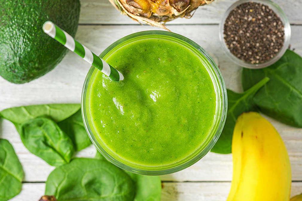 Zu sehen ist ein grüner Smoothie im Glas aus der Vogelperspektive auf einer Holzfläche. Drum herum sieht man im Anschnitt: eine Avocado, Spinatblätter, eine Banane, Ein Schälchen mit Chia-Samen, eine Ananas