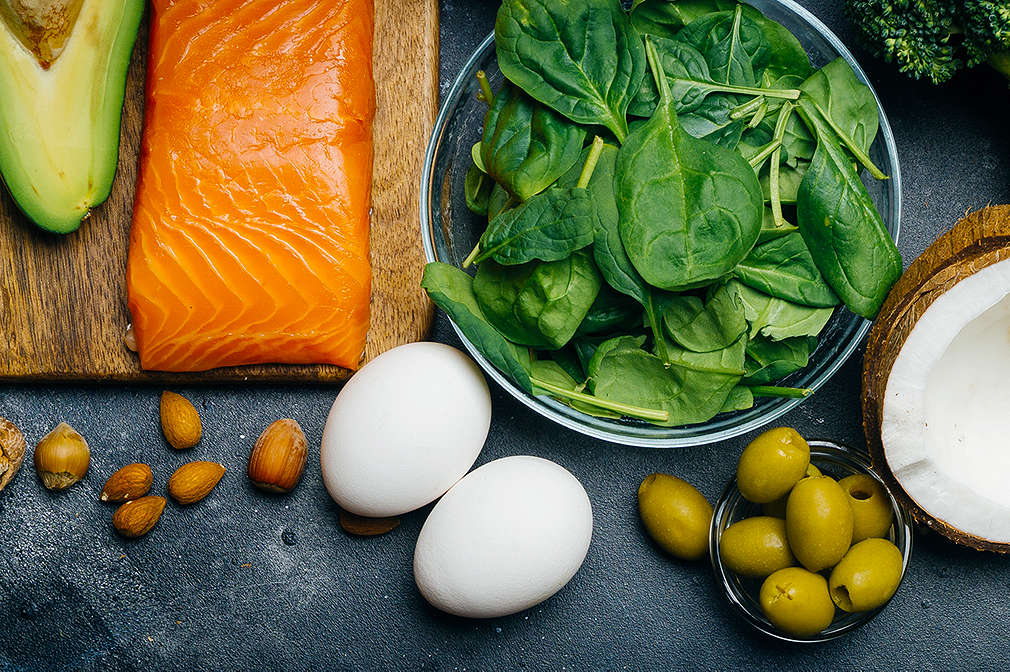 Verschiedene frische Lebensmittel wie Lachs, Käse, Eier, Nüsse, Kokosnuss, Oliven, Avocado und grünes Gemüse liegen auf einer dunklen Arbeitsfläche