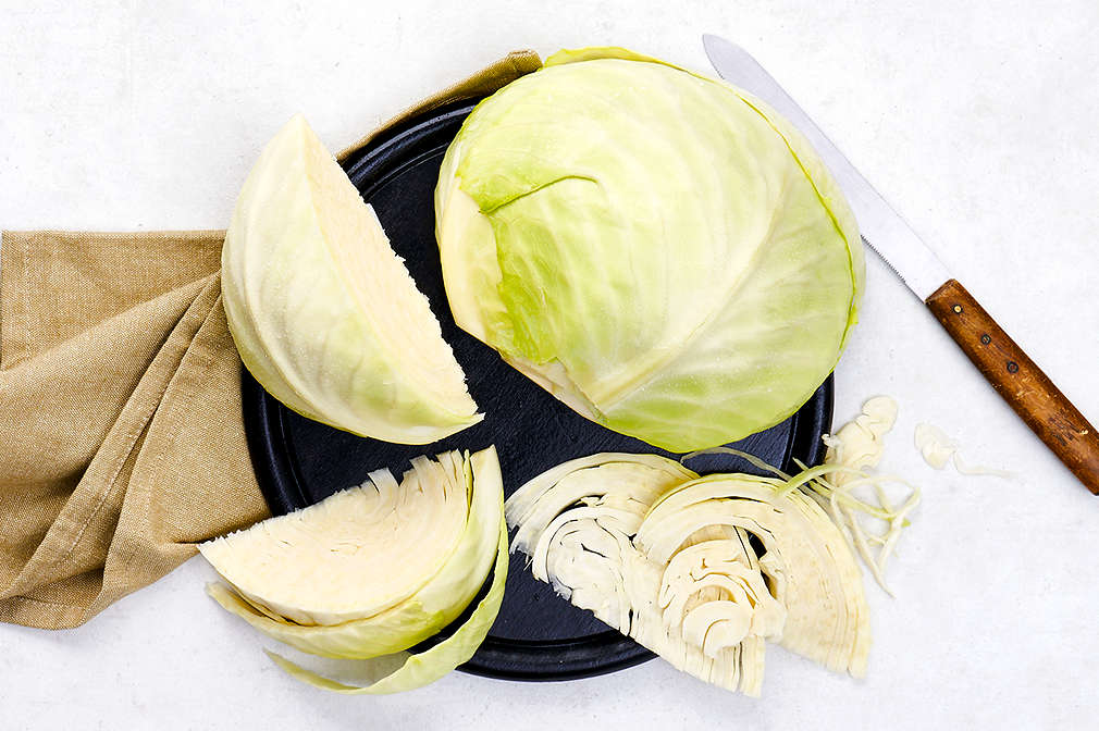 Heller Untergrund, links ein braunes Geschirrtuch, darüber ein runder flacher schwarzer Teller, auf dem eine Hälfte Weißkohl und drei weitere Spalten Weißkohl liegen; rechts vom Teller liegt ein Schneidemesser mit braunem Holzgriff