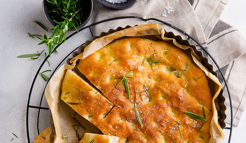 Zu sehen ist eine Focaccia in einer runden flachen Form auf einem runden Gitter, darunter ein weiß-graues Geschirrtuch, links über der Backform sind zwei Schälchen mit Rosmarin und grobem Salz.