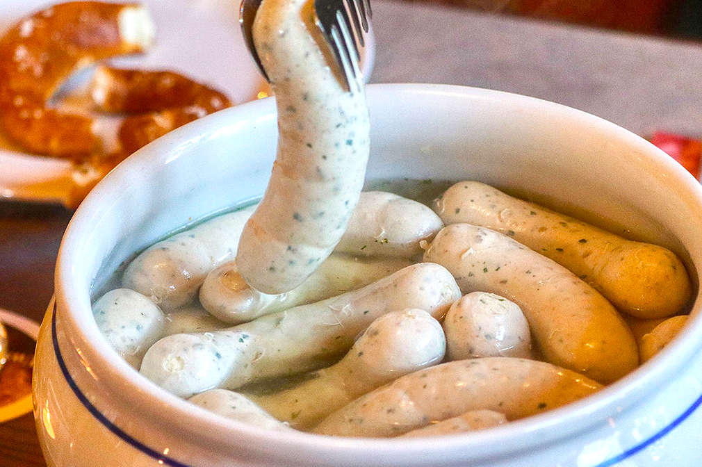 Weißwürste schwimmen in Kochwasser in einer weißen Keramikschüssel mit typisch bayerischem blau-weißem Muster; im Hintergrund erkennt man Brezeln