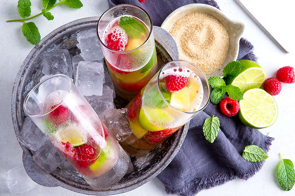 Drei Gläser in einem Eimer voller Eiswürfel,  gefüllt mit Himbeeren, Limetten, Minze und Orange. Im Hintergrund sieht man ein dunkelblaues Geschirrtuch, darauf ein Schälchen mit Rohrzucker, eine halbierte Limette, Minze und Himbeeren