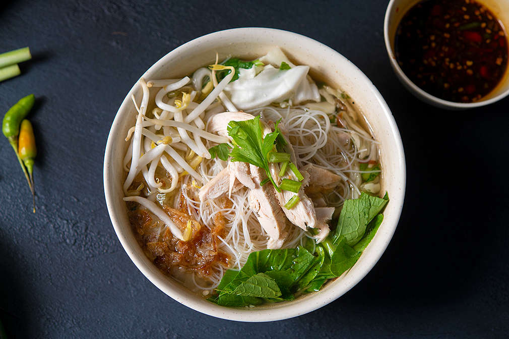 Ein Suppengericht mit Reisnudeln ist in einer Schale angerichtet, daneben steht Sojasauce