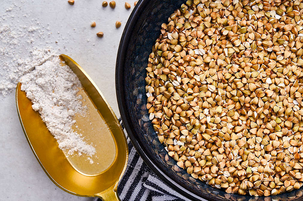 Auf einem goldenen Messlöffel ist etwas Mehl aus Buchweizensamen, daneben steht ein schwarzer Top mit Buchweizensamen drin