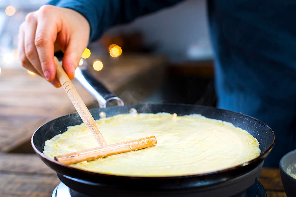 Crêpeteig wird mit Hilfe eines Teigverteilers dünn in der gusseisernen Pfanne verstrichen