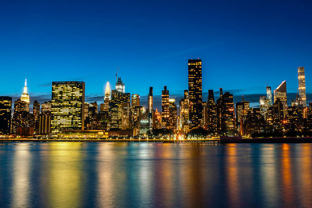 Zu sehen ist die New Yorker Skyline, die bei Nacht in tollen Gelb-, Rot-, Blau- und Lilatönen auf dem Wasser reflektiert