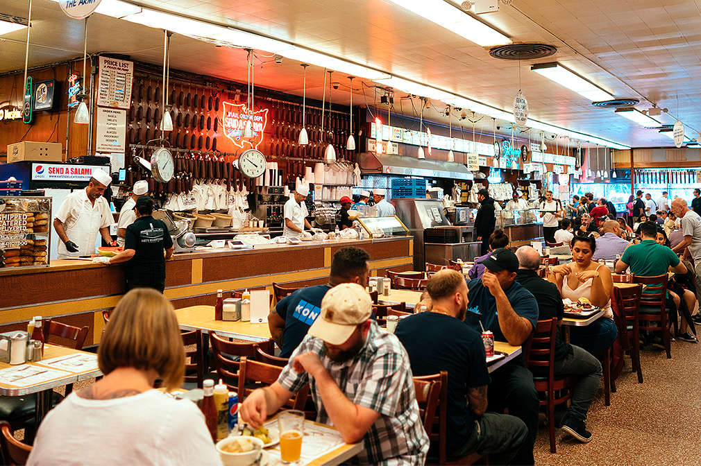 Innenansicht des Katz’s Deli in New York mit Bar und mehrreihigen Tischen mit Stühlen im Retro-Look, an denen teilweise Menschen sitzen und essen