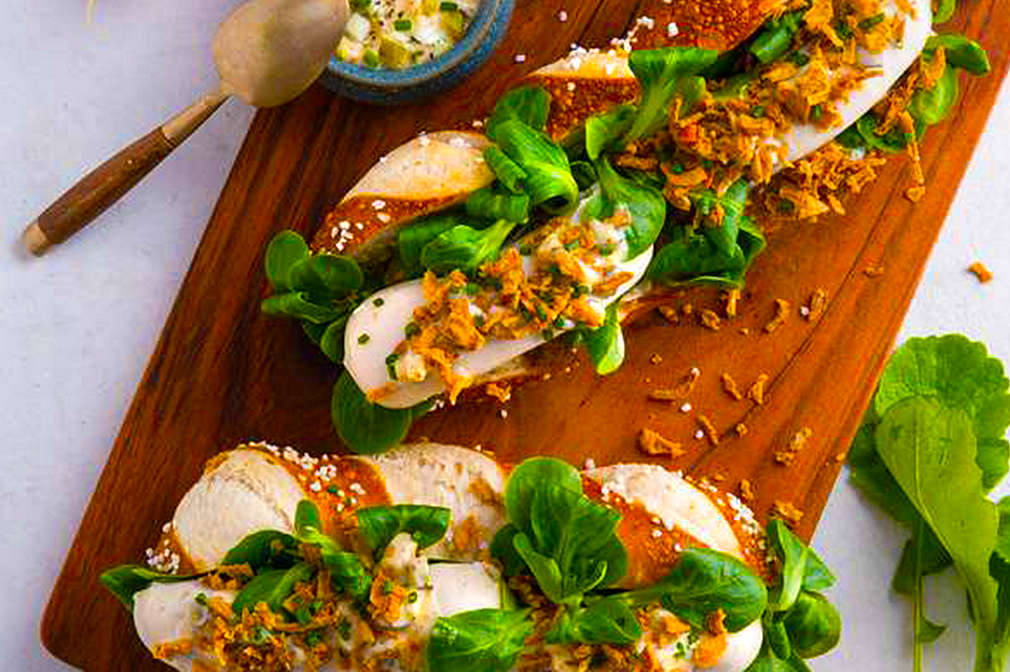 Ein hölzernes Schneidebrett mit zwei Hot Dogs in einer aufgeschnittenen Laugenstange mit Weißwurst, Feldsalat, Radieschen und einer kleinen Schale mit süßem Senf vor einem hellen Untergrund