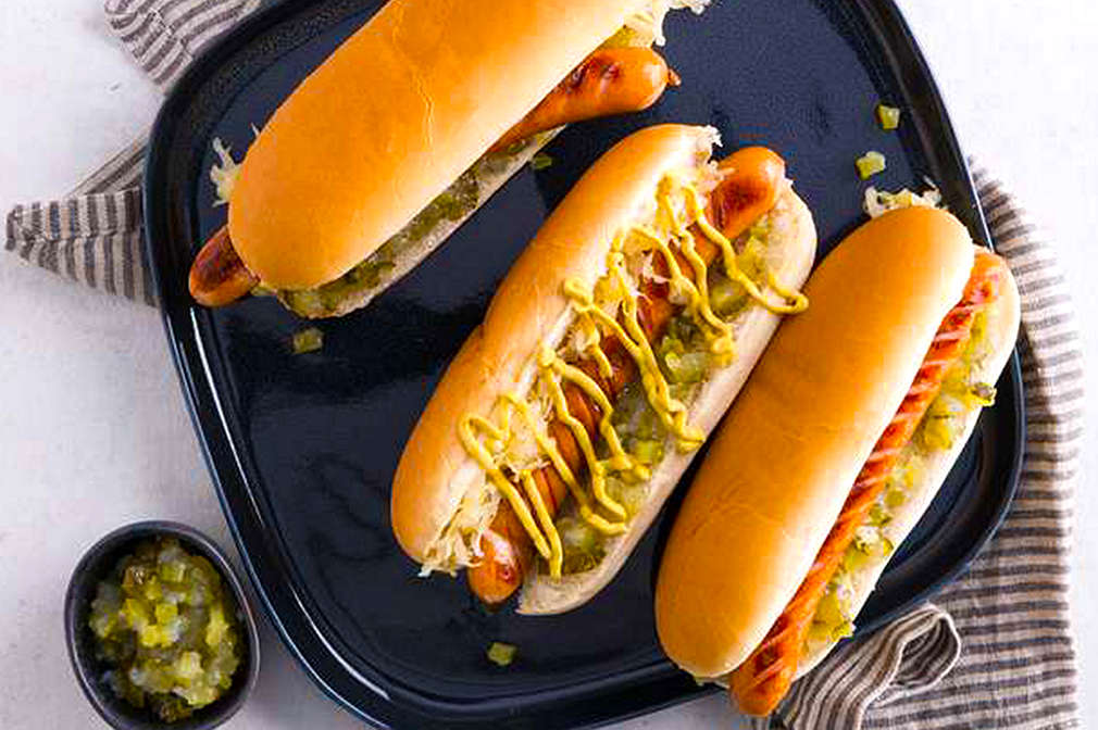 Drei Hot Dogs mit Frankfurter Würstchen, Gurken-Relish und Senf liegen auf einem großen schwarzen Teller neben einer kleinen Schale mit Relish vor einem hellen Untergrund