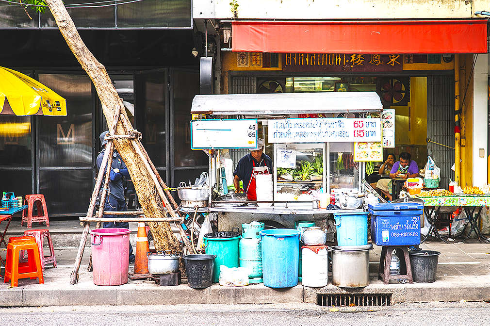 Zu sehen ist eine typische Garküche in den Straßen Bangkoks, wo Pad Thai frisch im Wok zubereitet und als Streetfood verkauft wird; neben dem Imbiss-Stand ist ein gelber Sonnenschirm mit einem Tisch und Hockern zum Sitzen erkennbar
