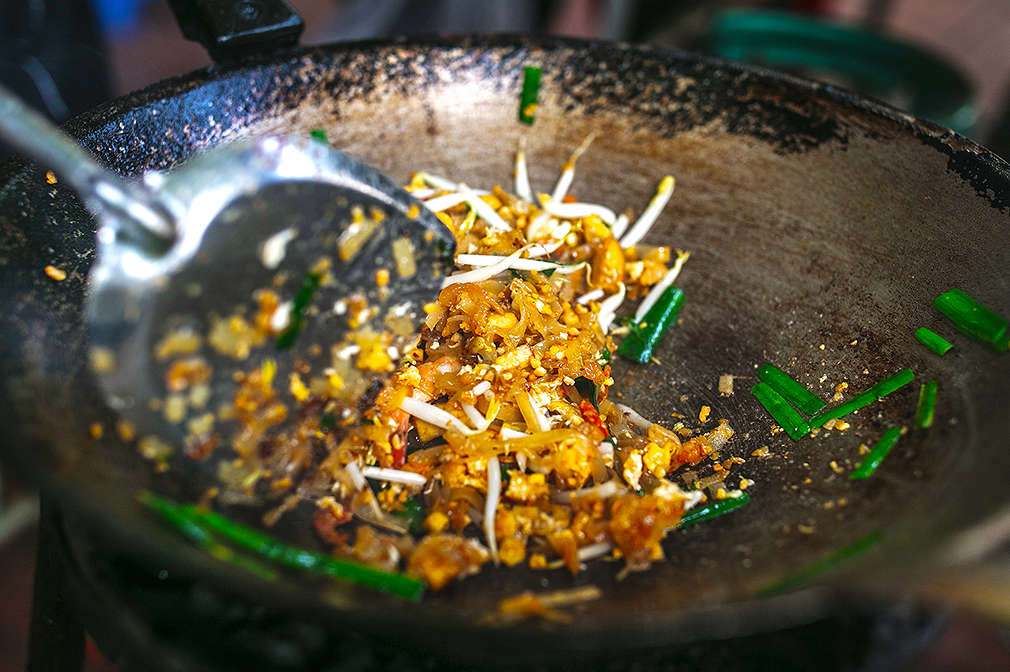 Ein Wok, in dem die frischen Pad-Thai-Zutaten gerade mit einem Pfannenwender verrührt werden, damit sich die Aromen vermischen