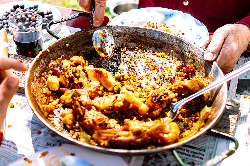 Eine Pfanne Paella Valenciana steht auf dem Tisch. Eine Person isst mit dem Löffel direkt aus der Pfanne