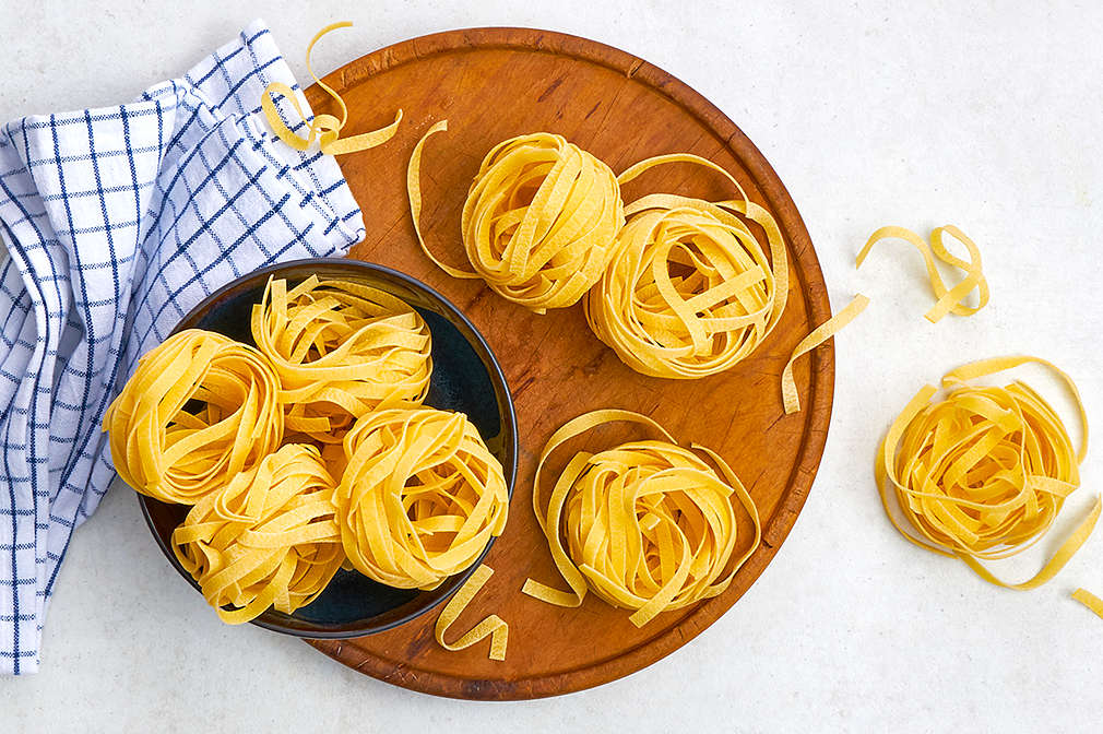 Auf einem runden Holzbrett steht eine schwarze Schüssel, sowohl auf dem Brett als auch in der Schüssel liegen kleine Nester ungekochter Tagliatelle, daneben liegt ein blau kariertes Geschirrtuch
