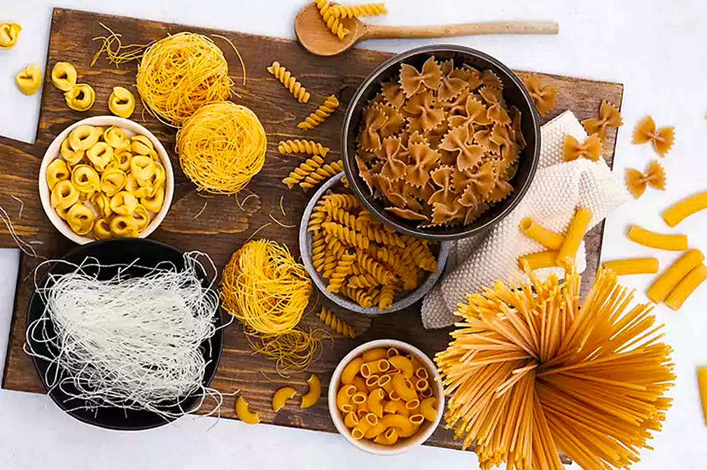 Verschiedene Nudelsorten liegen auf einem Holzbrett oder in Schüsseln nebeneinander, das Brett steht auf einem hellen Untergrund